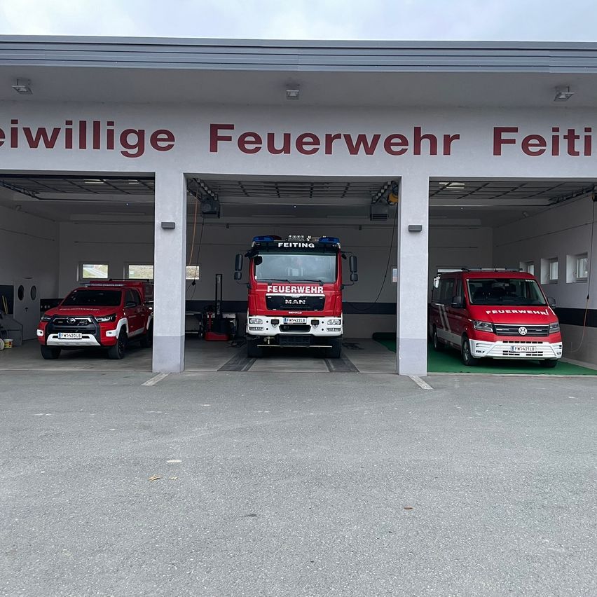 Eine Feuerwehrstation mit dem Namen Freiwillige Feuerwehr Feiti ist zu sehen. Drei rote Fahrzeuge sind drinnen geparkt, darunter ein LKW und zwei Autos.