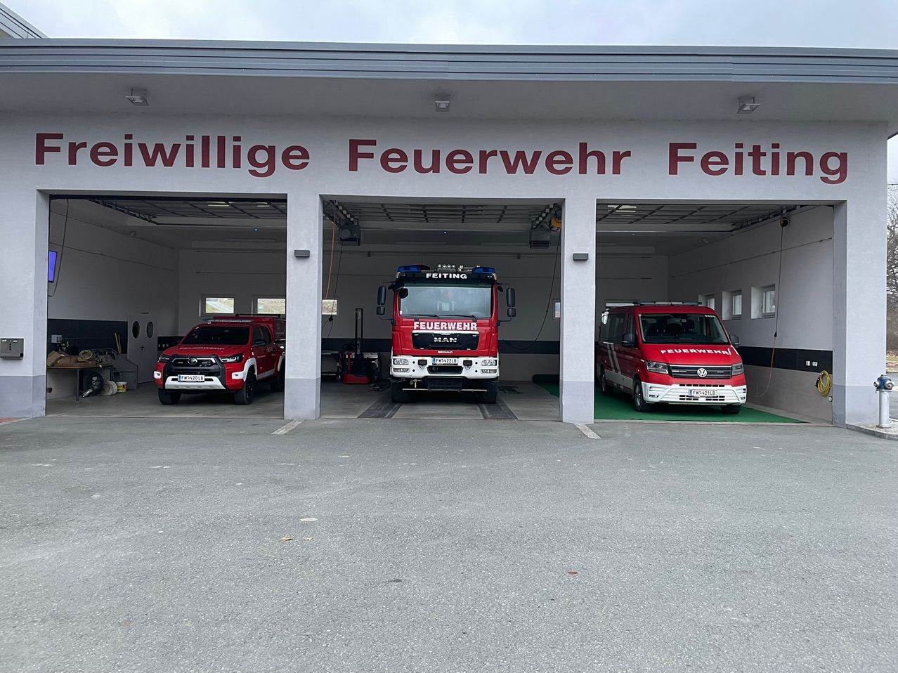 Eine Feuerwehrstation mit dem Namen Freiwillige Feuerwehr Feiti ist zu sehen. Drei rote Fahrzeuge sind drinnen geparkt, darunter ein LKW und zwei Autos.