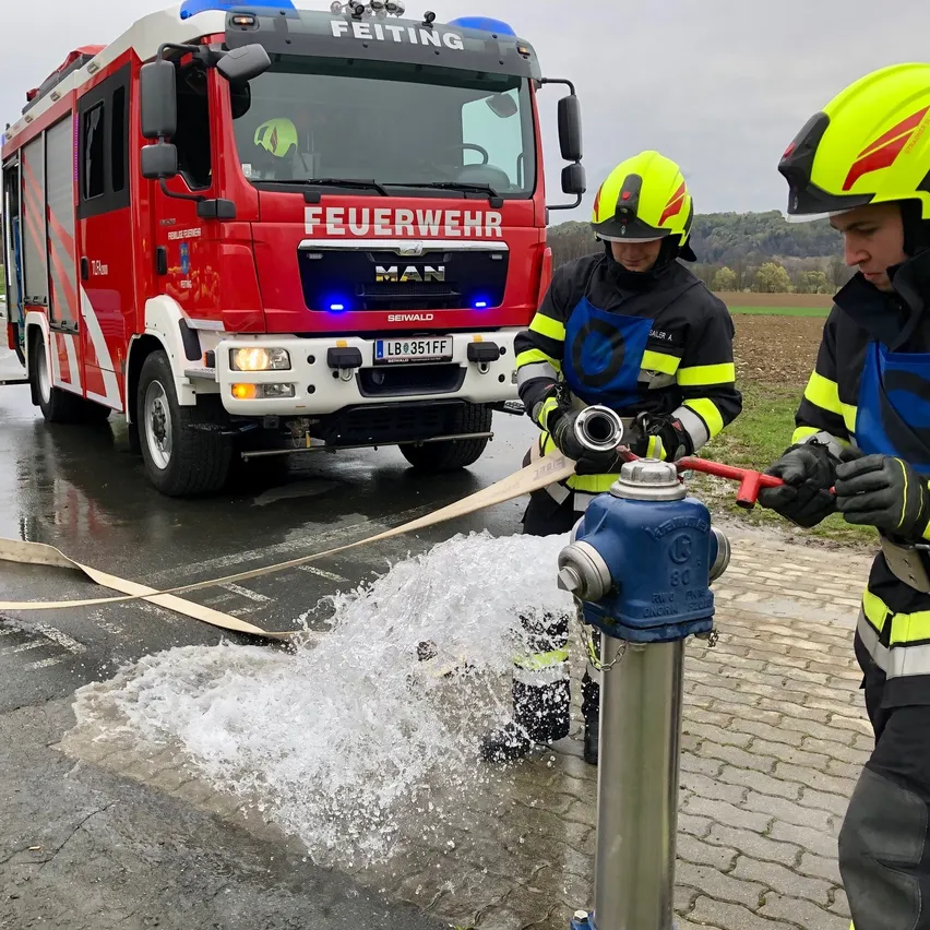 Zwei Feuerwehrleute öffnen an einem regnerischen Tag einen Hydranten mit einem roten Feuerwehrauto im Hintergrund.