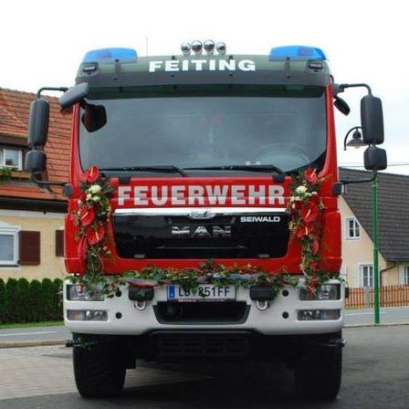 Ein roter Feuerwehrwagen mit 'Feuerwehr' und 'Feiting' auf der Front. Er hat Kränze und ein Nummernschild LB 851 FF. Dahinter steht ein Haus mit Zaun und Bäumen.