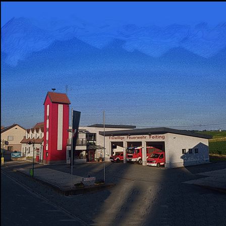 Eine Feuerwehr in Feiting zeigt einen roten Turm und Feuerwehrfahrzeuge. Es ist von einem grünen Feld und Häusern umgeben unter einem blauen Himmel.