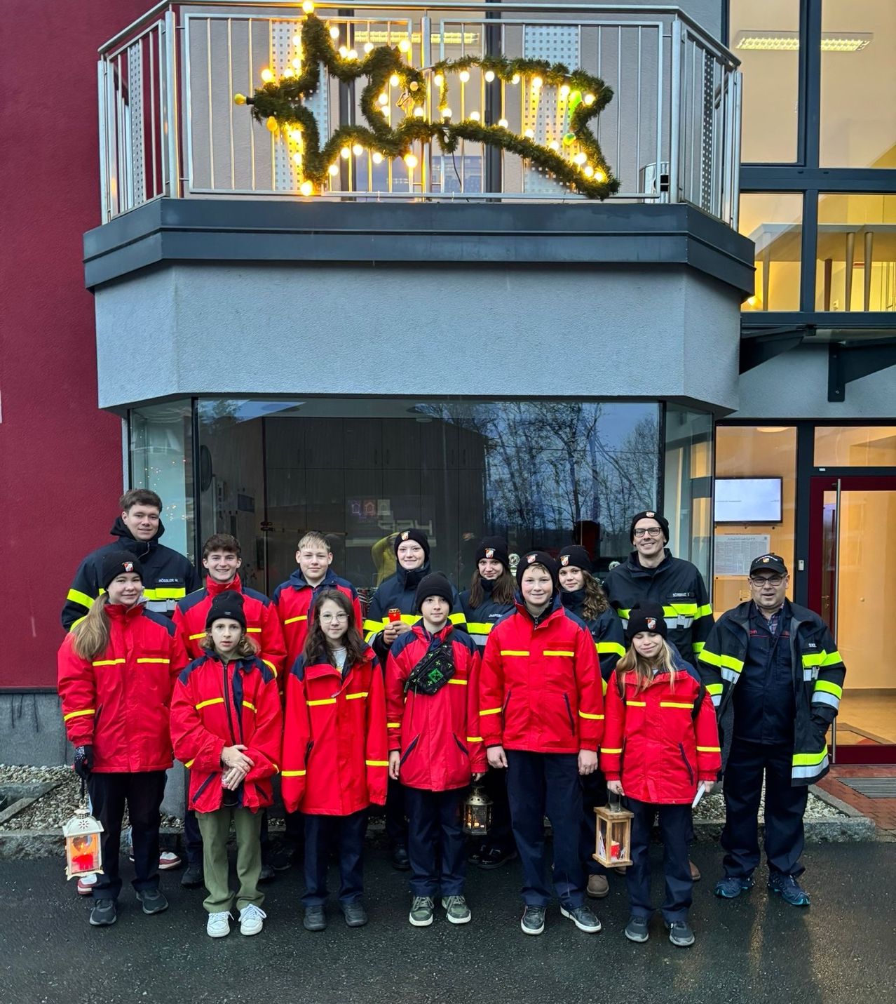 Eine Gruppe von Personen in roten Jacken mit gelben Streifen posiert für ein Foto vor einem Gebäude mit Balkon. Einige halten Laternen, und über dem Eingang befindet sich eine dekorative Girlande.