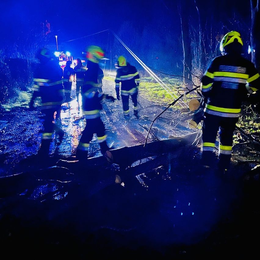 Feuerwehrleute in gelben und schwarzen Uniformen gehen bei Nacht auf einer nassen Straße. Sie tragen Helme und tragen Ausrüstung. Ein Baumstamm liegt auf der Straße.