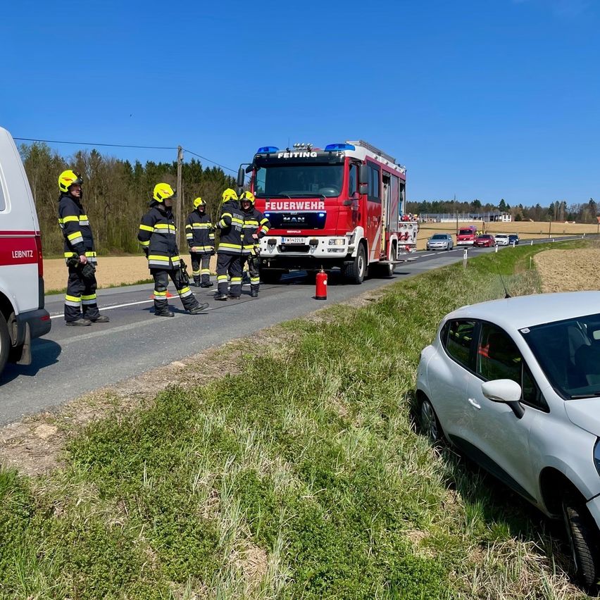 Feuerwehrleute in Uniform stehen neben einem Feuerwehrwagen und einem beschädigten Auto am Straßenrand. Der Feuerwehrwagen ist geparkt und mehrere Autos fahren vorbei.