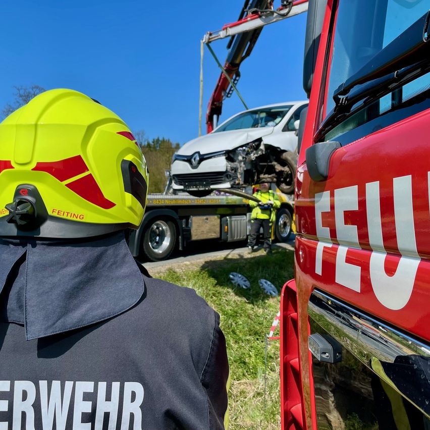 Ein Feuerwehrmann in einem gelben Helm beobachtet, wie ein Feuerwehrwagen ein beschädigtes weißes Auto anhebt. Ein anderer Arbeiter steht in der Nähe.