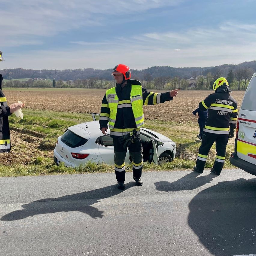 Drei Notfallsanitäter stehen neben einem weißen Auto, das von der Straße abgekommen ist. Einer zeigt auf das Fahrzeug, ein anderer hält eine Plastiktüte. Ein Feuerwehrwagen ist in der Nähe geparkt.