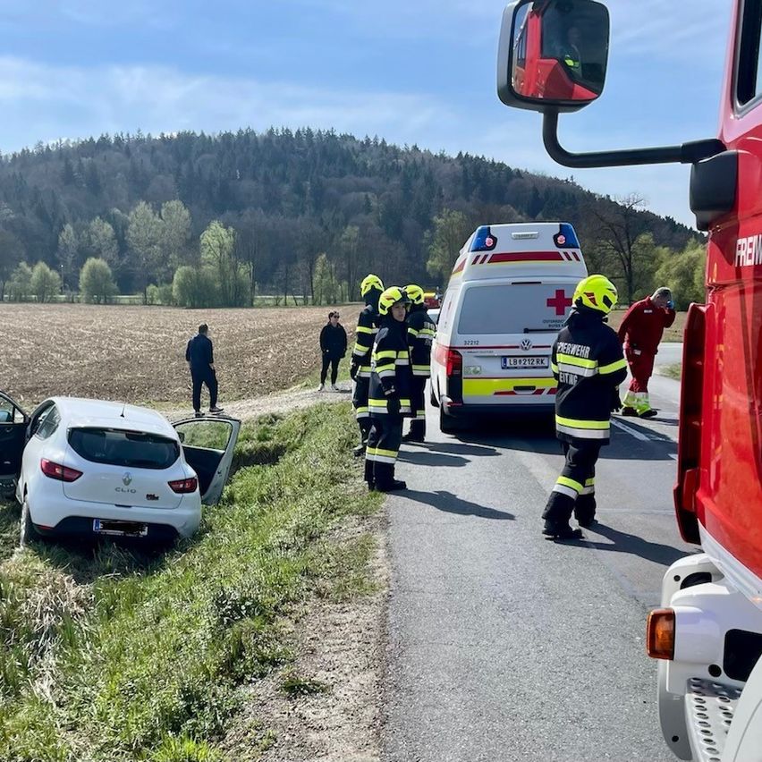 Rettungskräfte in gelben Helmen kümmern sich um einen Renault Clio, der von der Straße abgekommen ist. Ein Krankenwagen und ein Feuerwehrauto sind in der Nähe geparkt.