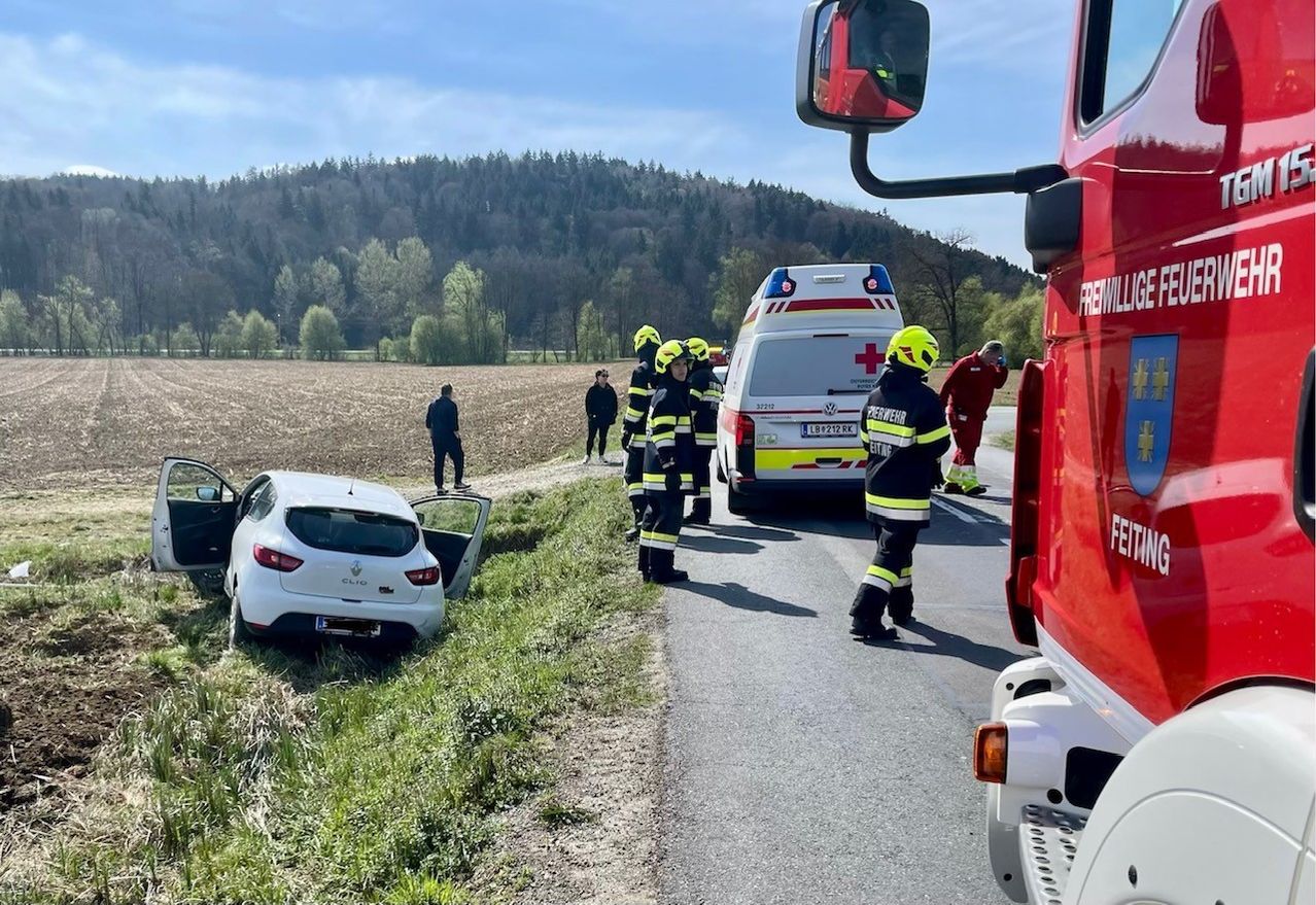 Rettungskräfte in gelben Helmen kümmern sich um einen Renault Clio, der von der Straße abgekommen ist. Ein Krankenwagen und ein Feuerwehrauto sind in der Nähe geparkt.