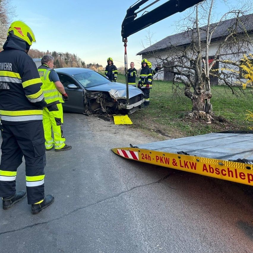 Rettungskräfte entfernen ein beschädigtes Auto von der Straßenseite. Ein Abschleppwagen hebt das Fahrzeug an, und mehrere Feuerwehrleute stehen in der Nähe.