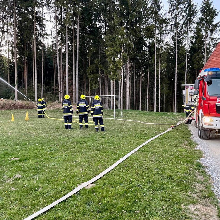 Feuerwehrleute üben auf einem Feld mit Feuerwehrauto und Schläuchen. Im Hintergrund steht ein Tor.