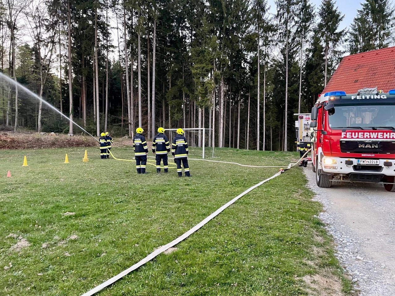 Feuerwehrleute üben auf einem Feld mit Feuerwehrauto und Schläuchen. Im Hintergrund steht ein Tor.