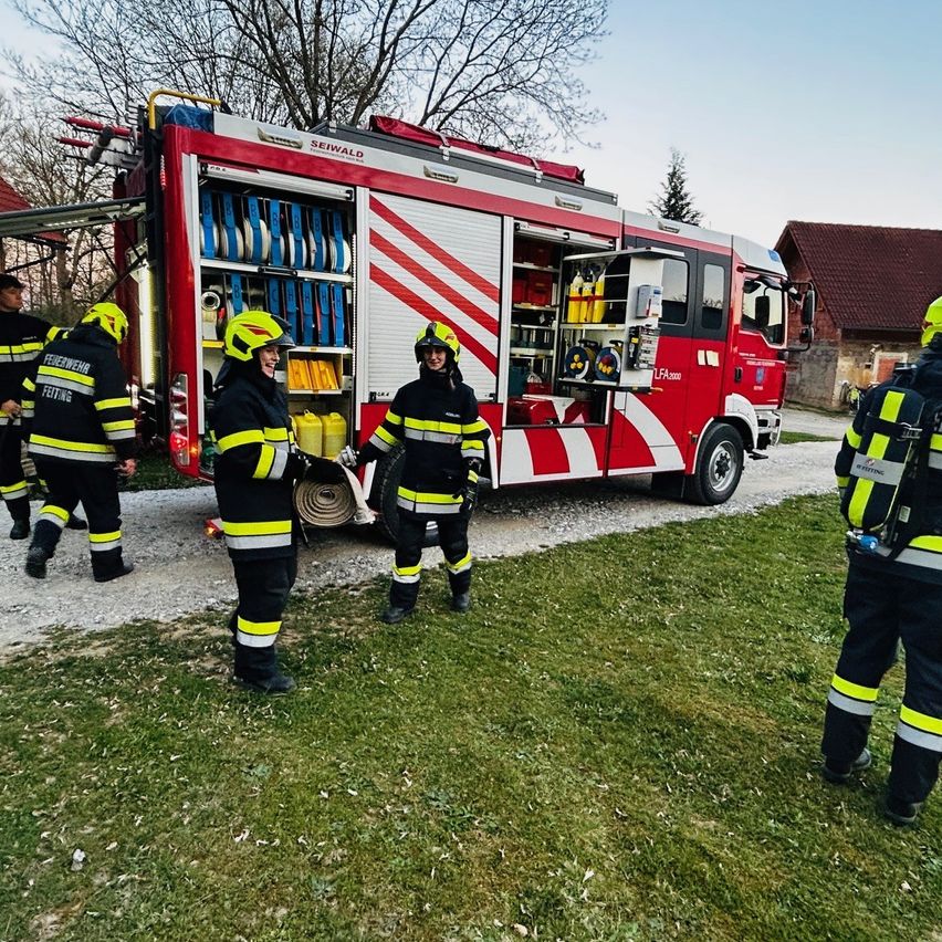 Eine Gruppe Feuerwehrleute steht bei einem roten Feuerwehrauto. Sie sind in Uniformen und Helmen gekleidet. In der Nähe befindet sich ein Haus und ein Baum.