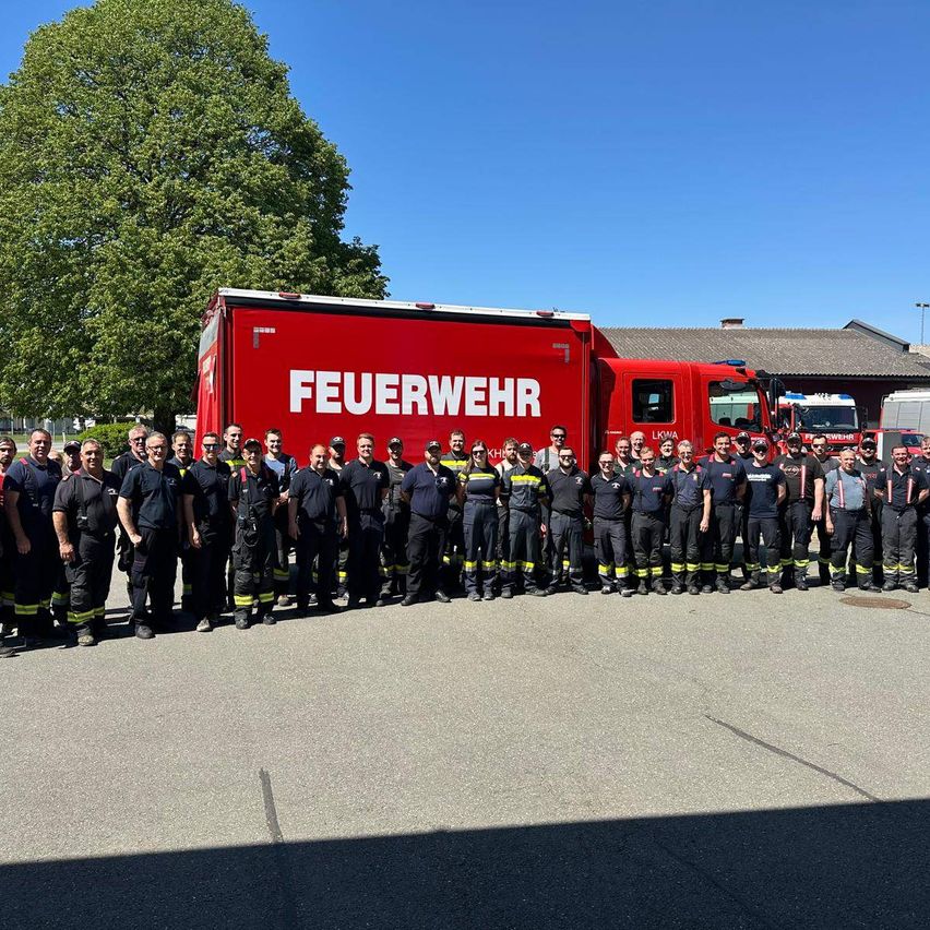 Eine Gruppe Feuerwehrleute steht vor einem roten Feuerwehrauto mit dem Wort 'FEUERWEHR' darauf. Sie sind alle in Uniform gekleidet.
