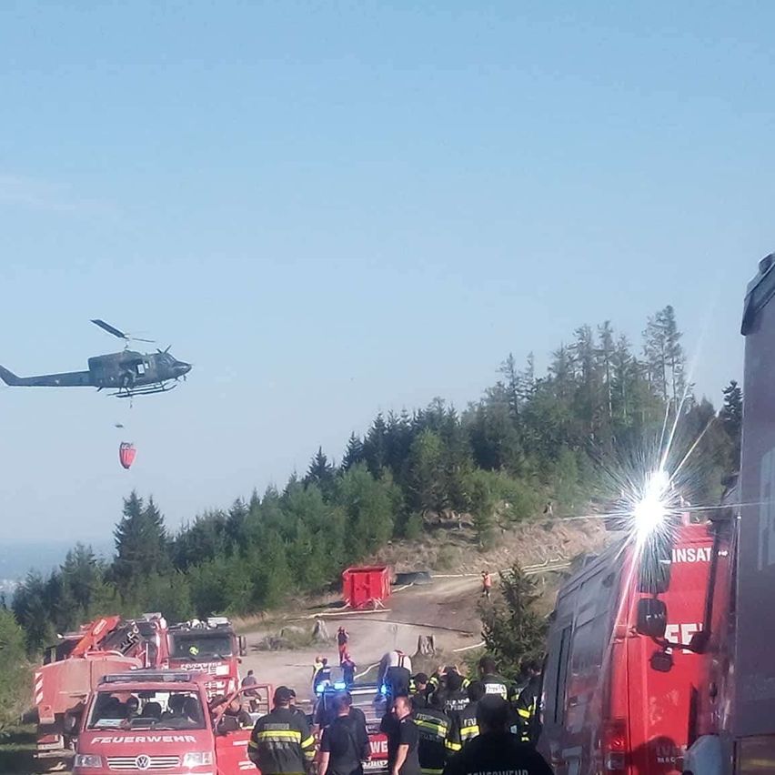 Ein Hubschrauber schwebt über einer Gruppe von Feuerwehrleuten und Rettungsfahrzeugen auf einem Berghang und lässt einen Rettungscontainer fallen.