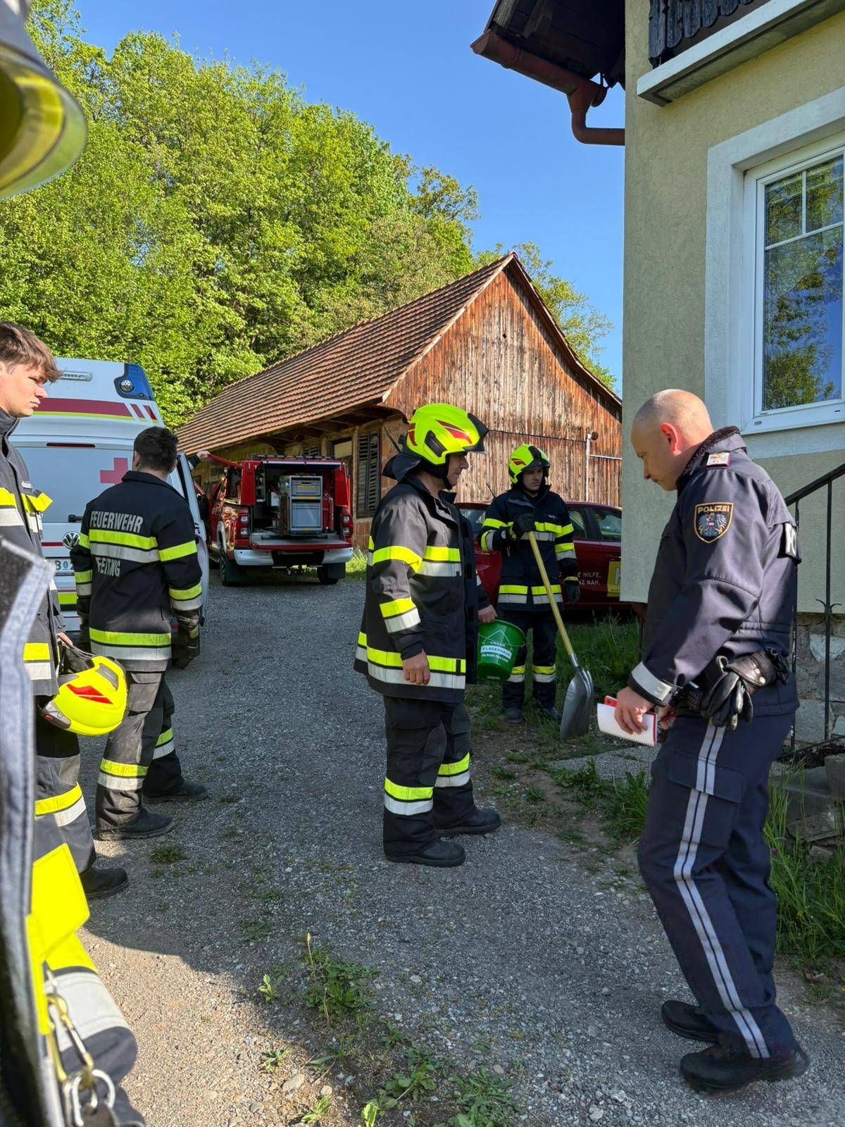 Feuerwehrleute und Polizeibeamte stehen vor einem Gebäude und besprechen möglicherweise eine Situation. Dahinter sind ein Feuerwehrwagen und ein Polizeiauto geparkt.