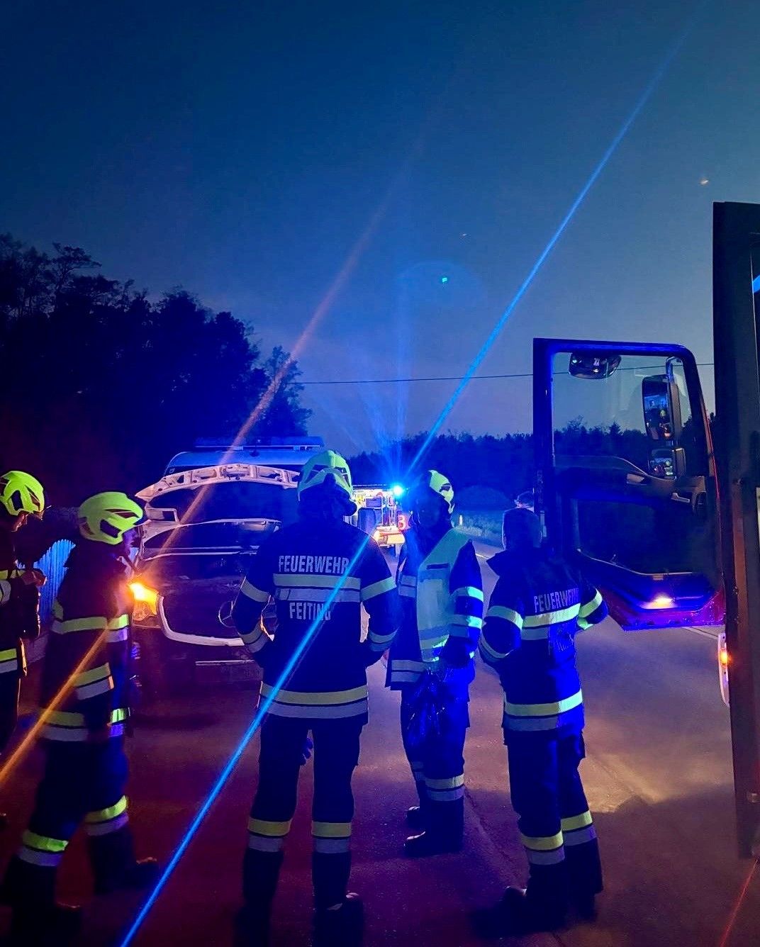 In der Nacht stehen Feuerwehrleute in der Nähe eines Autos mit offenem Motorhaube, einer hält eine Taschenlampe und der andere eine Taschenlampe.