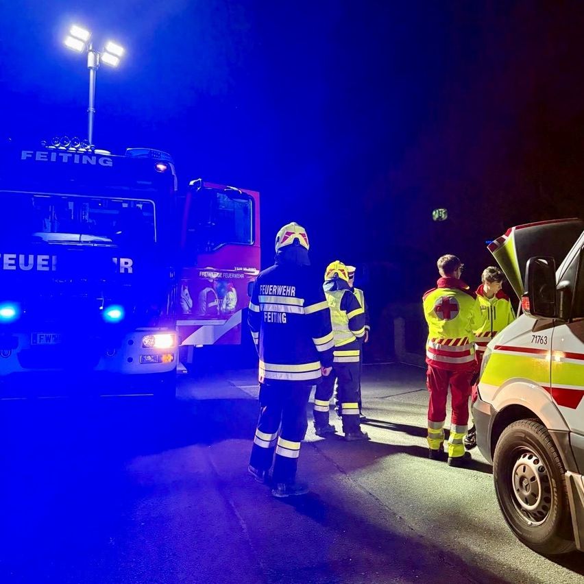 Feuerwehrleute in Uniformen stehen vor Rettungsfahrzeugen unter einem blauen Licht. Eine Person in rot-weißer Uniform spricht.