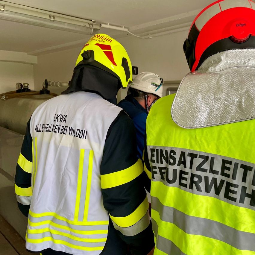 Drei Feuerwehrleute stehen in einem Raum und tragen Schutzwesten. Der Feuerwehrmann vorne hat einen gelben Helm mit der Aufschrift 'LKWA'. Ein anderer hat einen roten Helm. Sie überprüfen wahrscheinlich etwas im Raum.