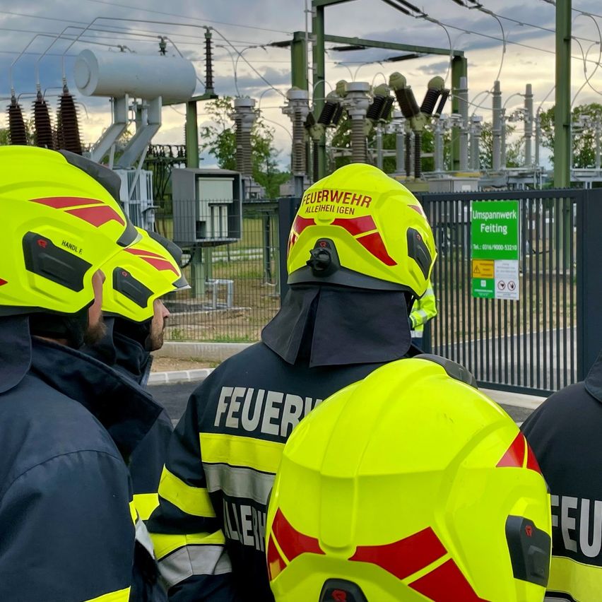 Feuerwehrleute in gelben Helmen stehen in der Nähe eines Kraftwerks und beobachten etwas vor sich. Im Hintergrund ist ein grünes Schild mit Text und Zahlen zu sehen.