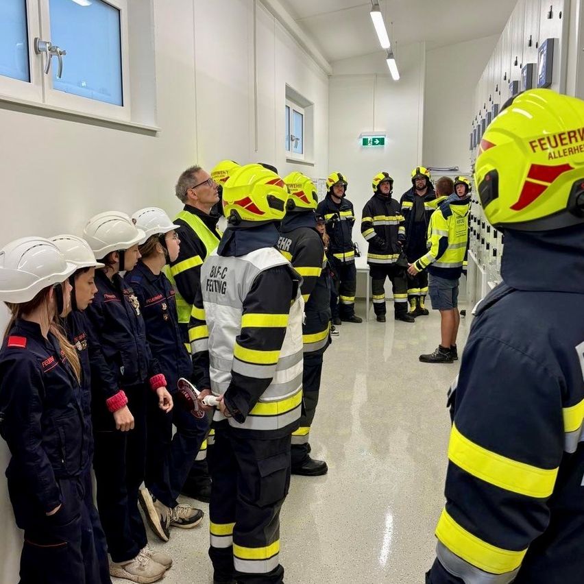 Eine Gruppe von Kindern mit weißen Helmen steht in einem Raum mit Feuerwehrleuten. Ein Feuerwehrmann hält einen Schuh, und andere stehen vor Schränken.