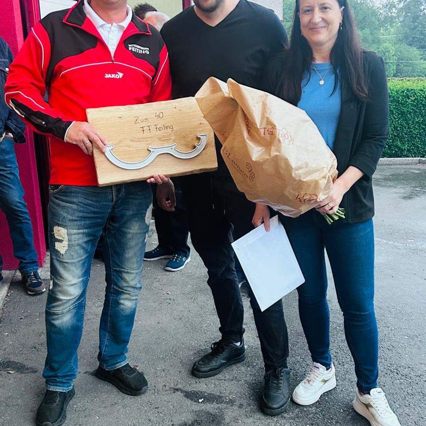 Drei Personen stehen zusammen draußen, eine hält eine Plakette, eine andere einen Papierbeutel und die dritte ein Papier. Eine Frau hält Blumen. Hinter ihnen steht ein rotes Gebäude und ein grüner Busch.