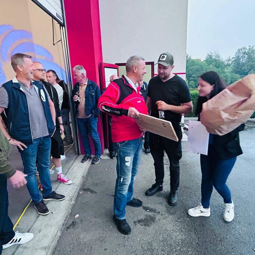 Männer in lässiger Kleidung versammeln sich vor einem Gebäude. Zwei Männer in der Mitte halten Ordner, während eine Frau einen braunen Papierbeutel hält. Bäume und ein Himmel sind im Hintergrund.