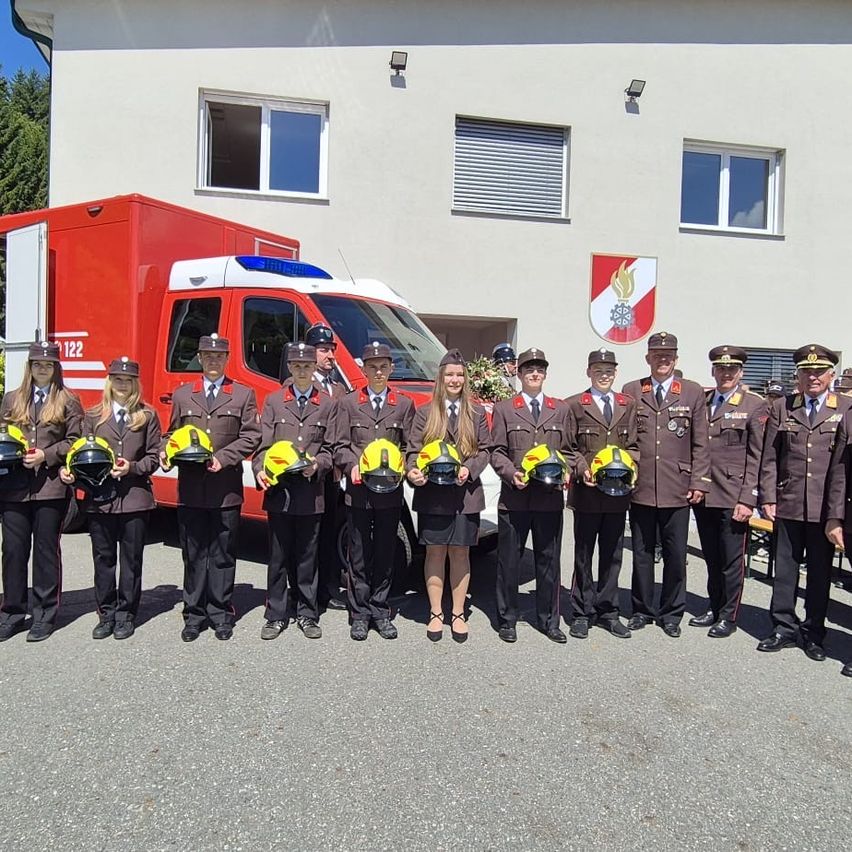 Eine Gruppe uniformierter Personen, wahrscheinlich Feuerwehrleute, steht vor einem Gebäude mit einem Krankenwagen und einem Logo und hält Helme.
