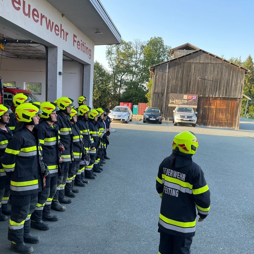 Eine Gruppe von Feuerwehrleuten in gelben Helmen und Uniformen steht in einer Reihe vor einer Feuerwache. Ein Kind in einem gelben Helm schaut zu.