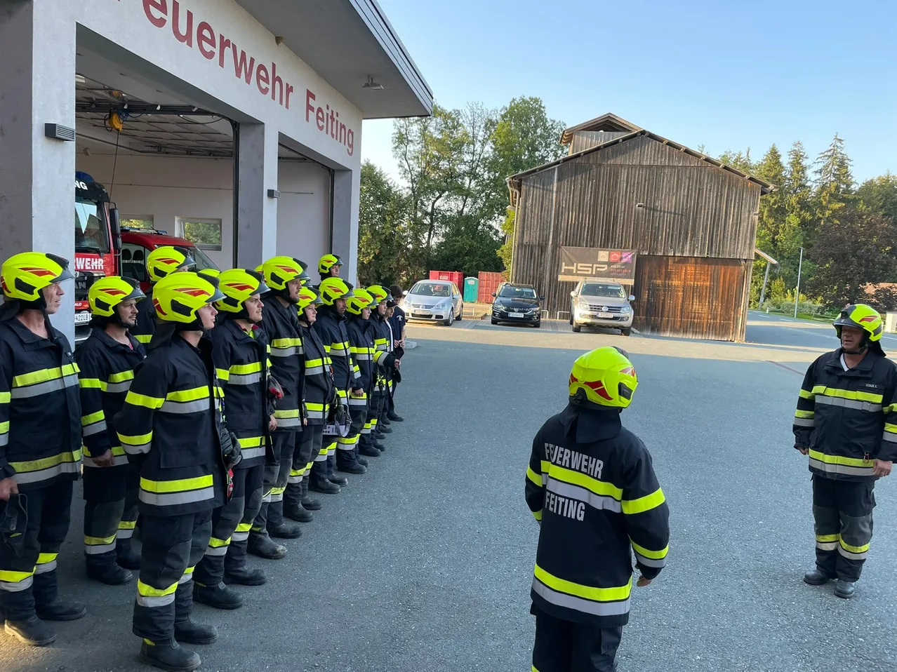 Eine Gruppe von Feuerwehrleuten in gelben Helmen und Uniformen steht in einer Reihe vor einer Feuerwache. Ein Kind in einem gelben Helm schaut zu.