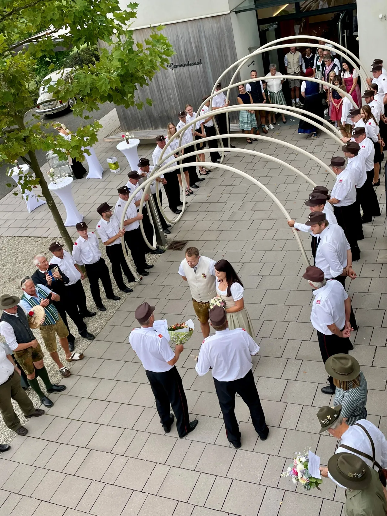 Eine Hochzeitszeremonie mit einem Paar in der Mitte, umgeben von Menschen in traditioneller Kleidung, die große weiße Bögen halten.