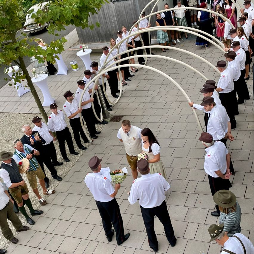 Eine Hochzeitszeremonie mit einem Paar in der Mitte, umgeben von Menschen in traditioneller Kleidung, die große weiße Bögen halten.