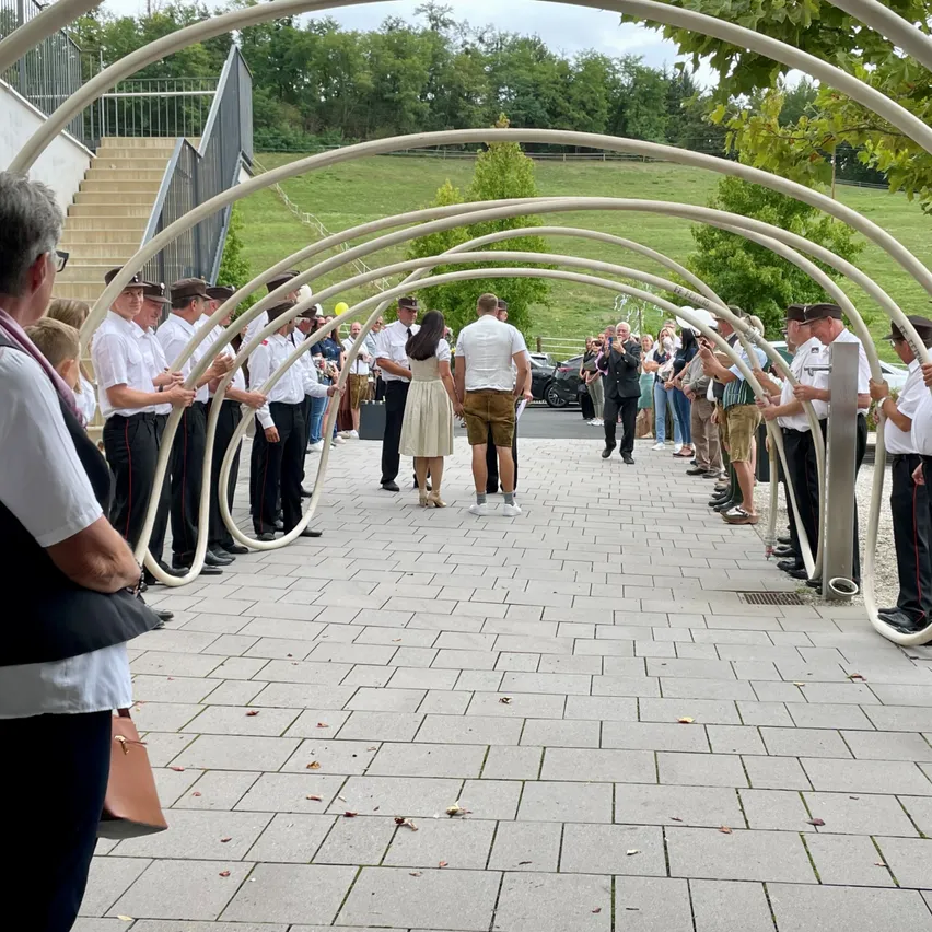 Eine Gruppe von Menschen in Uniformen und Hüten steht unter einer geschwungenen Struktur, möglicherweise für eine Zeremonie, mit Zuschauern in der Nähe.