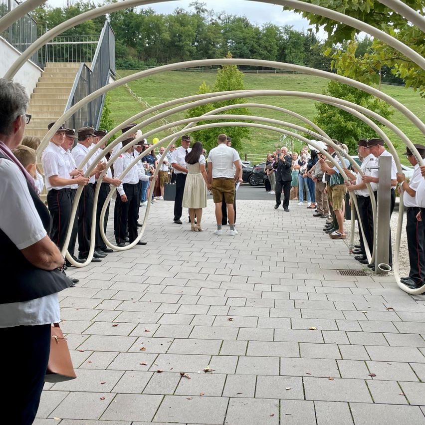 Eine Gruppe von Menschen in Uniformen und Hüten steht unter einer geschwungenen Struktur, möglicherweise für eine Zeremonie, mit Zuschauern in der Nähe.