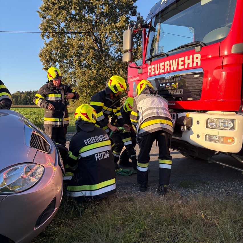 Feuerwehrleute in gelben und schwarzen Uniformen arbeiten an einem Verkehrsunfall am Straßenrand. Ein roter Feuerwehrwagen ist in der Nähe geparkt.