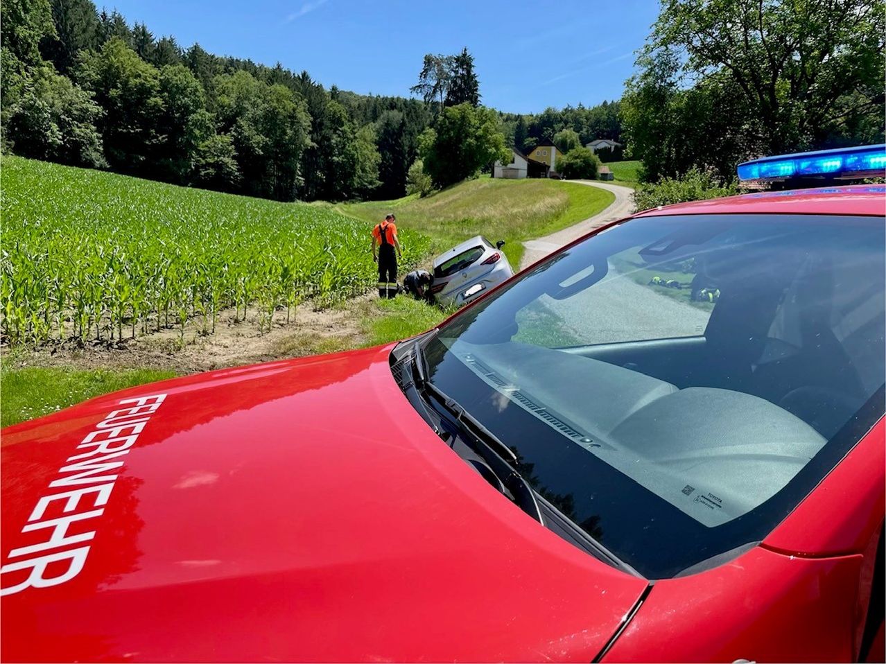 Ein roter Feuerwehrwagen und ein umgekipptes Auto neben einem Maisfeld. Eine Person in Orange überprüft das Auto, während eine andere kniet.