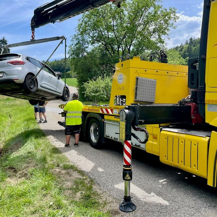 Ein silberner Wagen wird von einem Kran gehoben und von einem gelben Lastwagen geschleppt. Ein Mann in einer gelben Weste steht daneben.