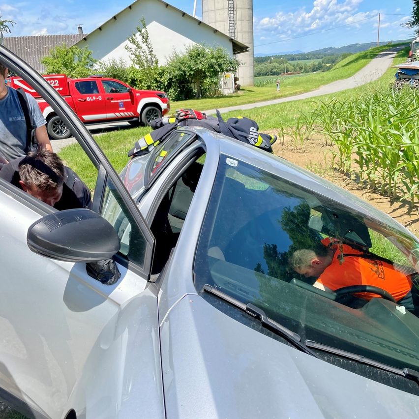 Zwei Personen helfen einem Fahrer in einem silbernen Auto, mit einem roten Feuerwehrauto im Hintergrund.