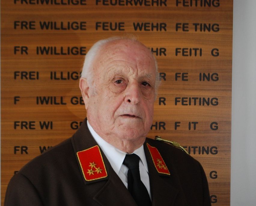 Ein älterer Mann in Uniform mit roten Schulterklappen steht vor einem hölzernen Schild. Er sieht ernst aus.
