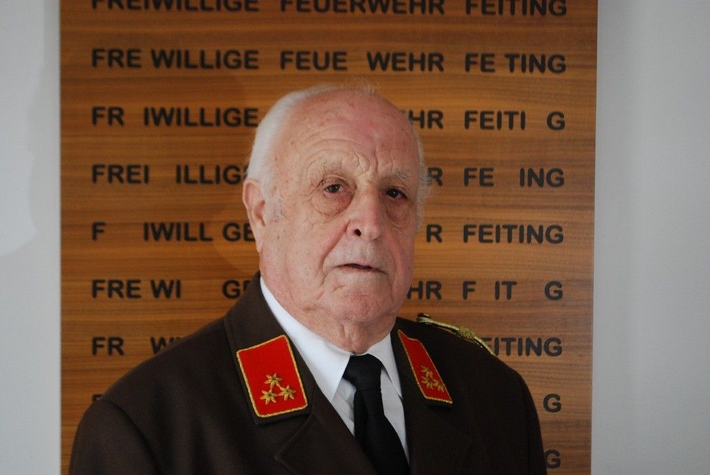 Ein älterer Mann in Uniform mit roten Schulterklappen steht vor einem hölzernen Schild. Er sieht ernst aus.