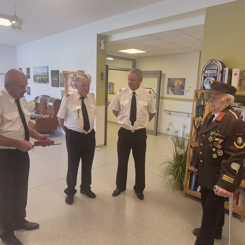 Drei Männer in Uniform stehen drinnen, einer hält ein Buch. Hinter ihnen steht ein dekorierter Mann in Uniform und ein Bücherregal voller Bücher und ein Jukebox.