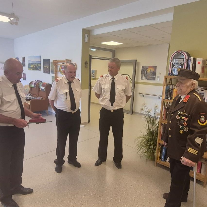 Drei Männer in Uniform stehen drinnen, einer hält ein Buch. Hinter ihnen steht ein dekorierter Mann in Uniform und ein Bücherregal voller Bücher und ein Jukebox.