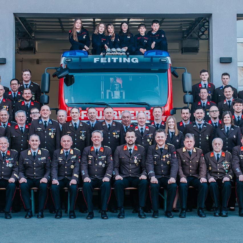 Eine Gruppe uniformierter Personen posiert vor einem Feuerwehrauto für ein Foto, wobei einige obenauf stehen.