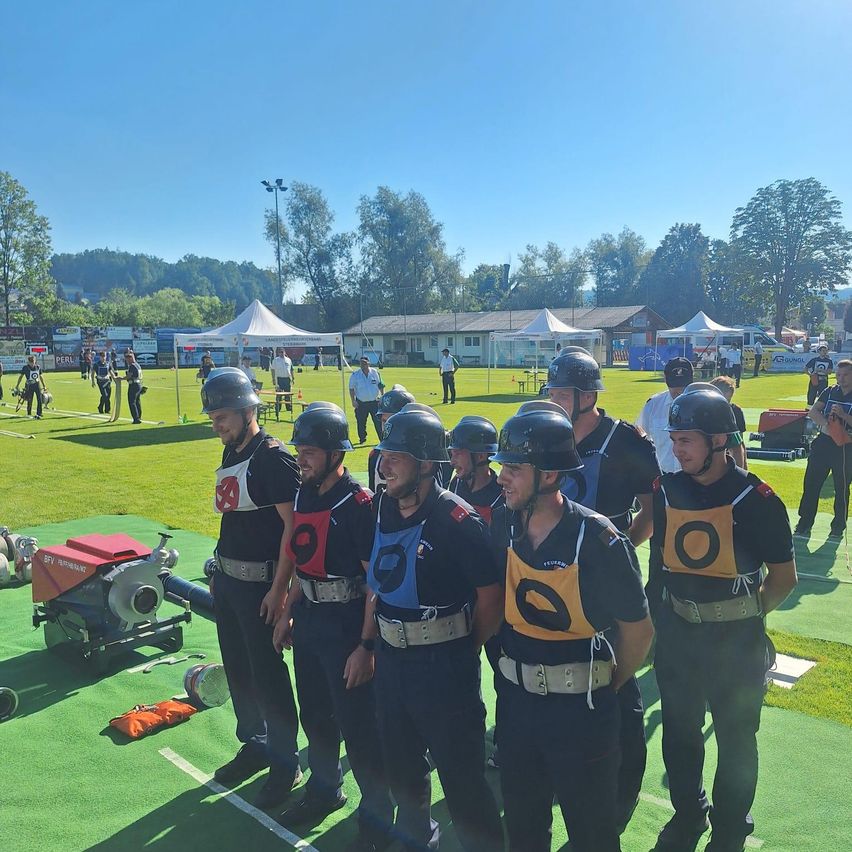 Gruppe von Personen in identischen Uniformen steht auf einem Sportplatz, möglicherweise in Vorbereitung auf einen Wettkampf. Bäume und Gebäude im Hintergrund.