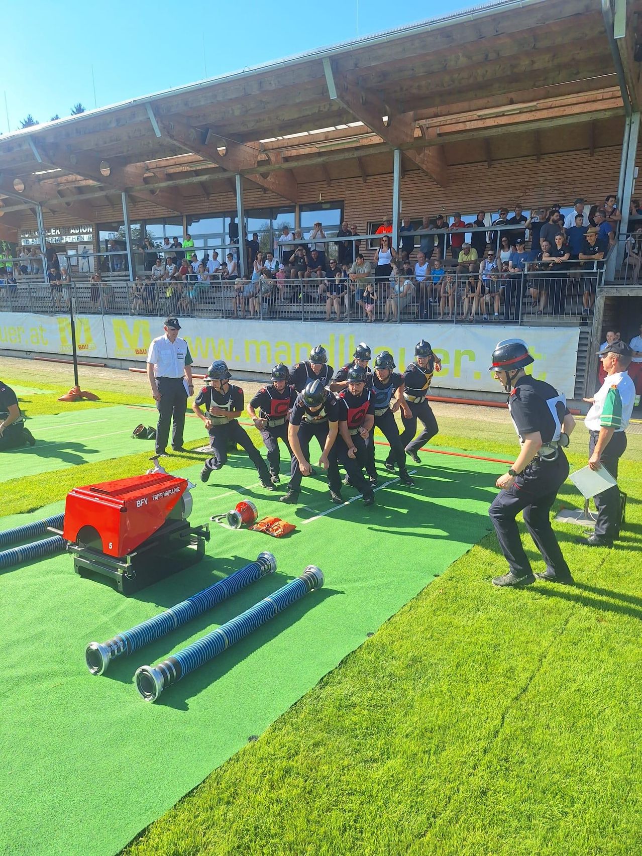 Eine Gruppe von Menschen in Helmen und Uniformen nimmt an einem Rennen auf einem grünen Feld teil. Zuschauer beobachten von den Tribünen aus. Ein Mann mit Papier und ein anderer Mann, der geht, sind im Vordergrund.