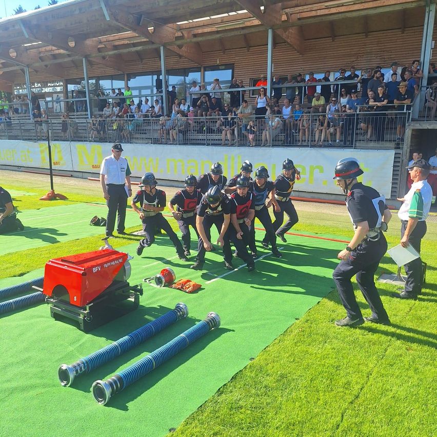 Eine Gruppe von Menschen in Helmen und Uniformen nimmt an einem Rennen auf einem grünen Feld teil. Zuschauer beobachten von den Tribünen aus. Ein Mann mit Papier und ein anderer Mann, der geht, sind im Vordergrund.