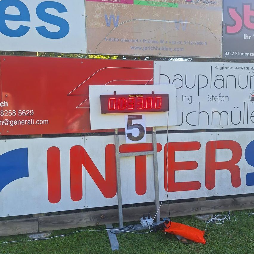 Eine digitale Außenuhr zeigt die Zeit 00:53:80 an. Sie steht vor einem Schild mit INTERS in roten und blauen Buchstaben. Weitere Schilder und Banner sind ebenfalls an der Wand befestigt.