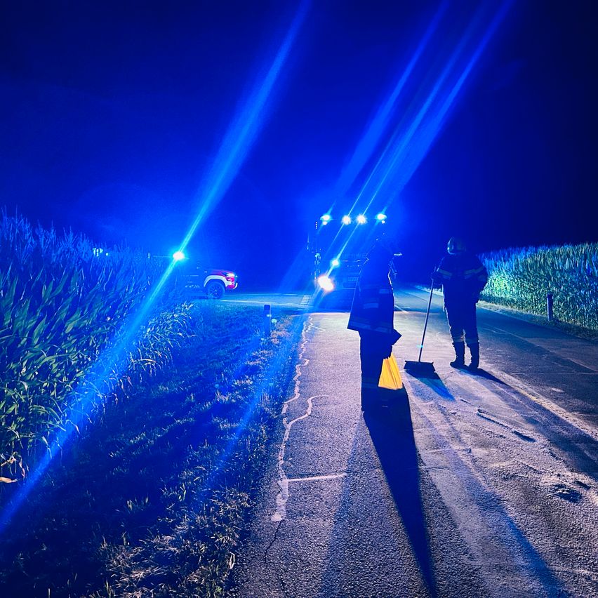 Zwei Feuerwehrleute stehen neben einem Fahrzeug mit Lichtern, das die Straße nachts fegt. Blaue Lichter beleuchten die Szene.