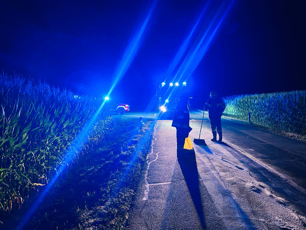 Zwei Feuerwehrleute stehen neben einem Fahrzeug mit Lichtern, das die Straße nachts fegt. Blaue Lichter beleuchten die Szene.