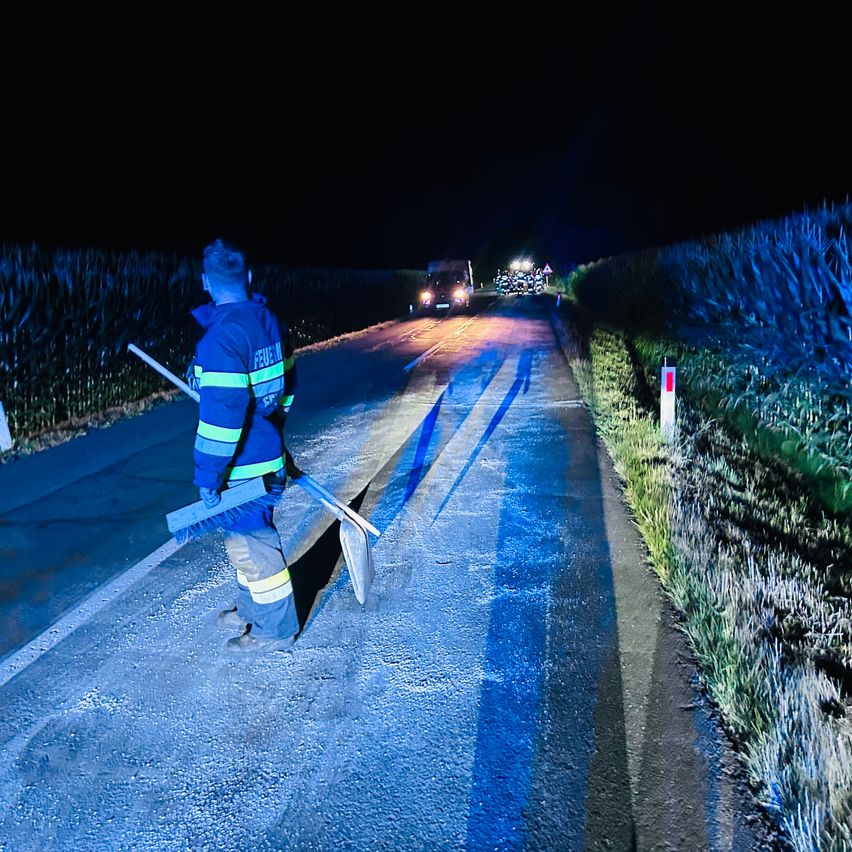 Ein Feuerwehrmann steht bei Nacht auf einer Landstraße und hält einen Besen und ein Werkzeug in der Hand, während ein Auto in der Ferne näherkommt.
