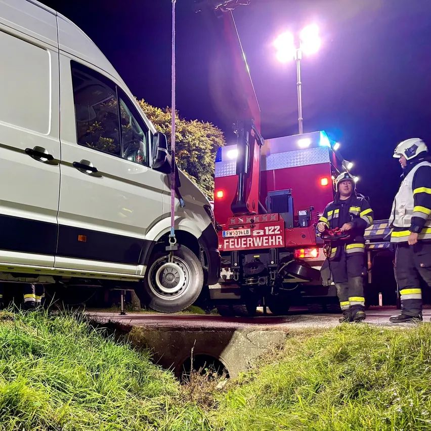 Ein weißer Lieferwagen steckt in einem Loch am Straßenrand. Ein Feuerwehrwagen steht dahinter mit eingeschalteten Lichtern. Vier Feuerwehrleute stehen um den Wagen herum, zwei von ihnen tragen Helme.